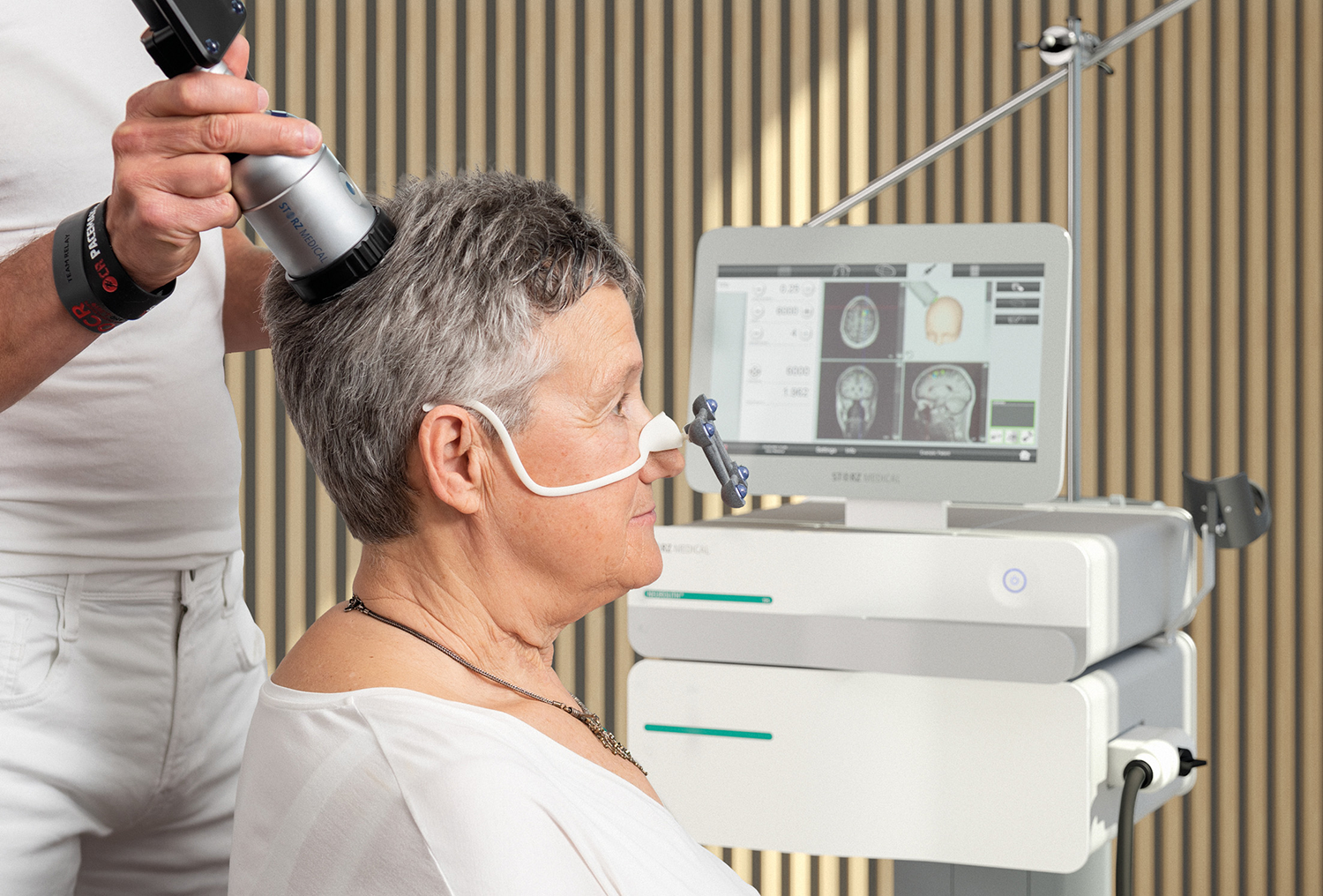Ältere Frau sitzt vor Gerät zur Behandlung von Alzheimer Demenz und Arzt hält Teil des Gerätes an den Kopf